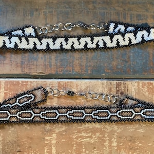 Peut inclure: Deux colliers ras du cou perlés avec fermoirs à chaîne argentés. L'un présente un motif en zigzag blanc et noir, l'autre un motif de maillons de chaîne marron et blanc, tous deux avec des bordures noires. Les colliers sont présentés sur une surface en bois.