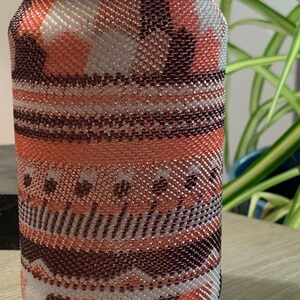May include: A decorative jar with a gold lid, covered in a beaded pattern. The beadwork features a mix of colors including white, peach, copper, and dark brown, creating a textured, patterned design. The jar sits on a wooden surface.
