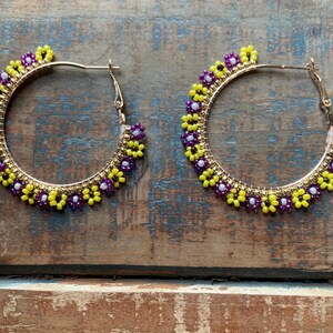 Puede incluir: Un par de pendientes de aro dorados con cuentas de flores de color morado y amarillo.