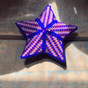 May include: A handcrafted, beaded star in shades of blue, pink, and red. The star is five-pointed, with a detailed woven pattern. The beads create a textured surface, and the star is set against a rustic wooden background.