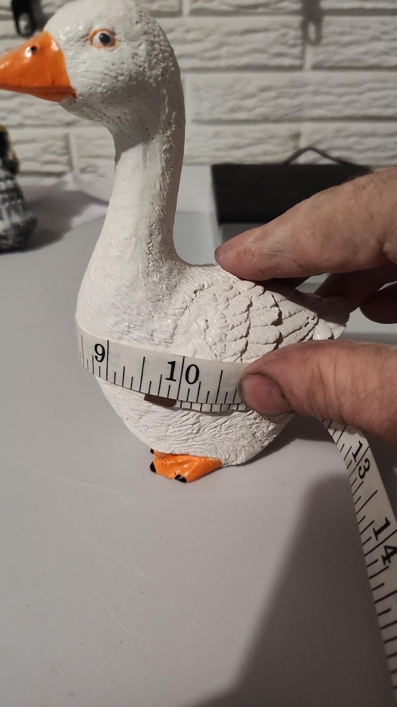May include: A white goose figurine with orange beak and feet, measured with a tape measure, showing a circumference of 10 inches.