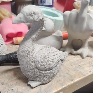 May include: A gray concrete goose figurine with detailed feathers. The goose is sitting on a wooden surface.
