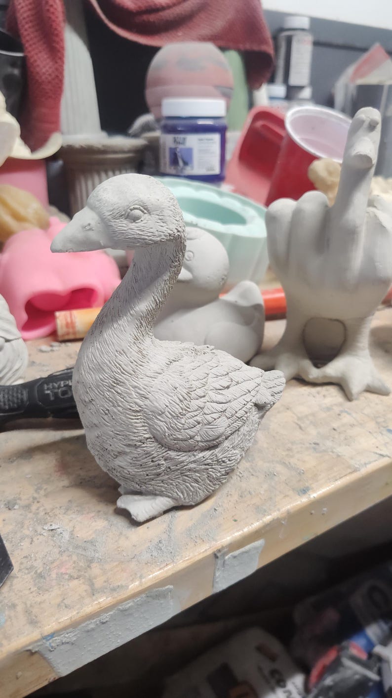 May include: A gray concrete goose figurine with detailed feathers. The goose is sitting on a wooden surface.