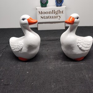 May include: Two white ceramic goose figurines with orange beaks and feet. The geese are facing each other. The box behind the geese says "Moonlight Statuary".