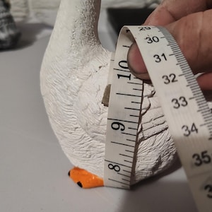 May include: White ceramic goose figurine with orange beak and feet. The goose is being measured with a tape measure, showing a measurement of 9 inches.