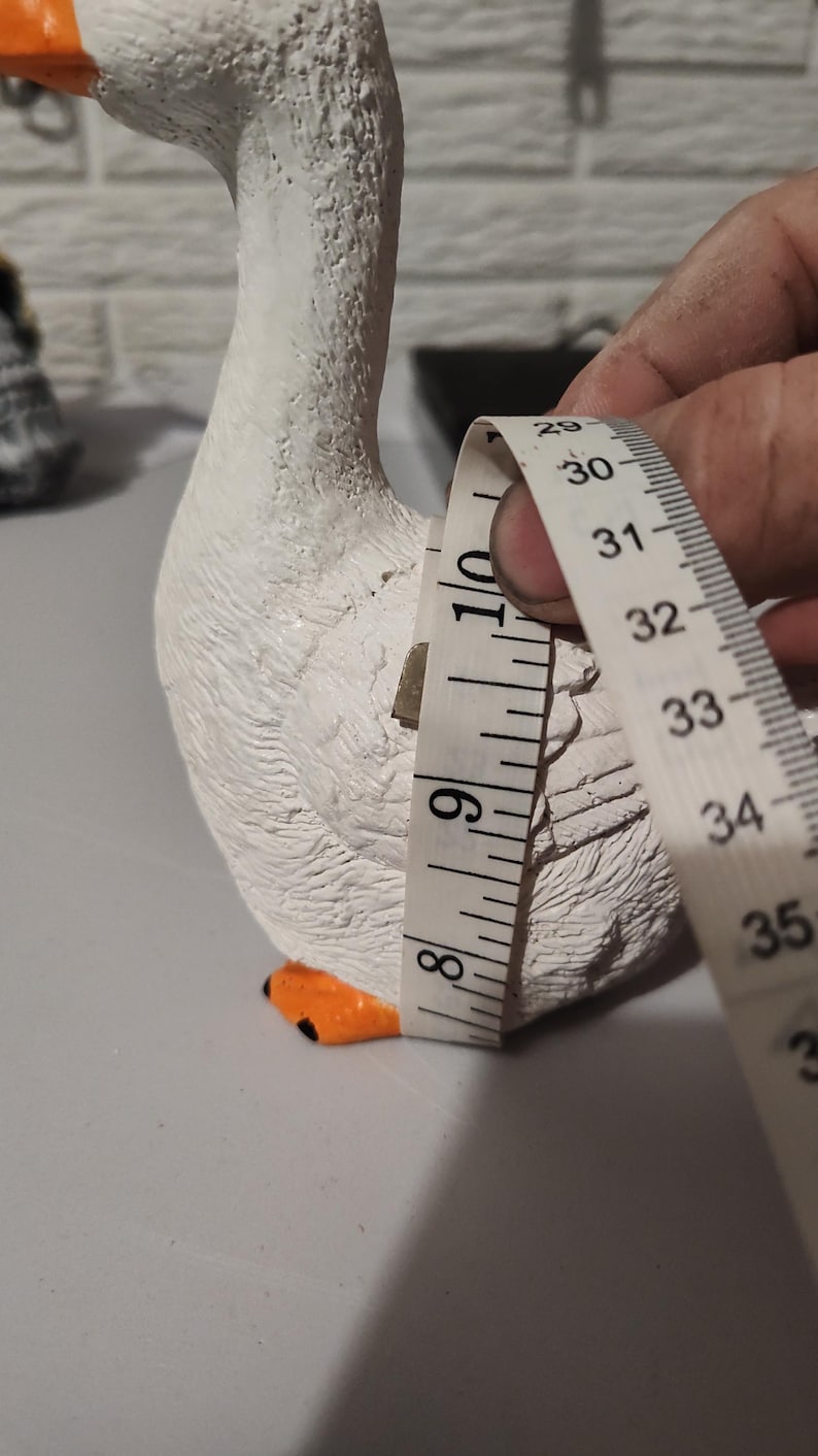 May include: White ceramic goose figurine with orange beak and feet. The goose is being measured with a tape measure, showing a measurement of 9 inches.