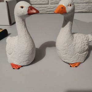 May include: Two white ceramic goose figurines with orange beaks and feet. The geese are facing each other and are positioned on a white surface.
