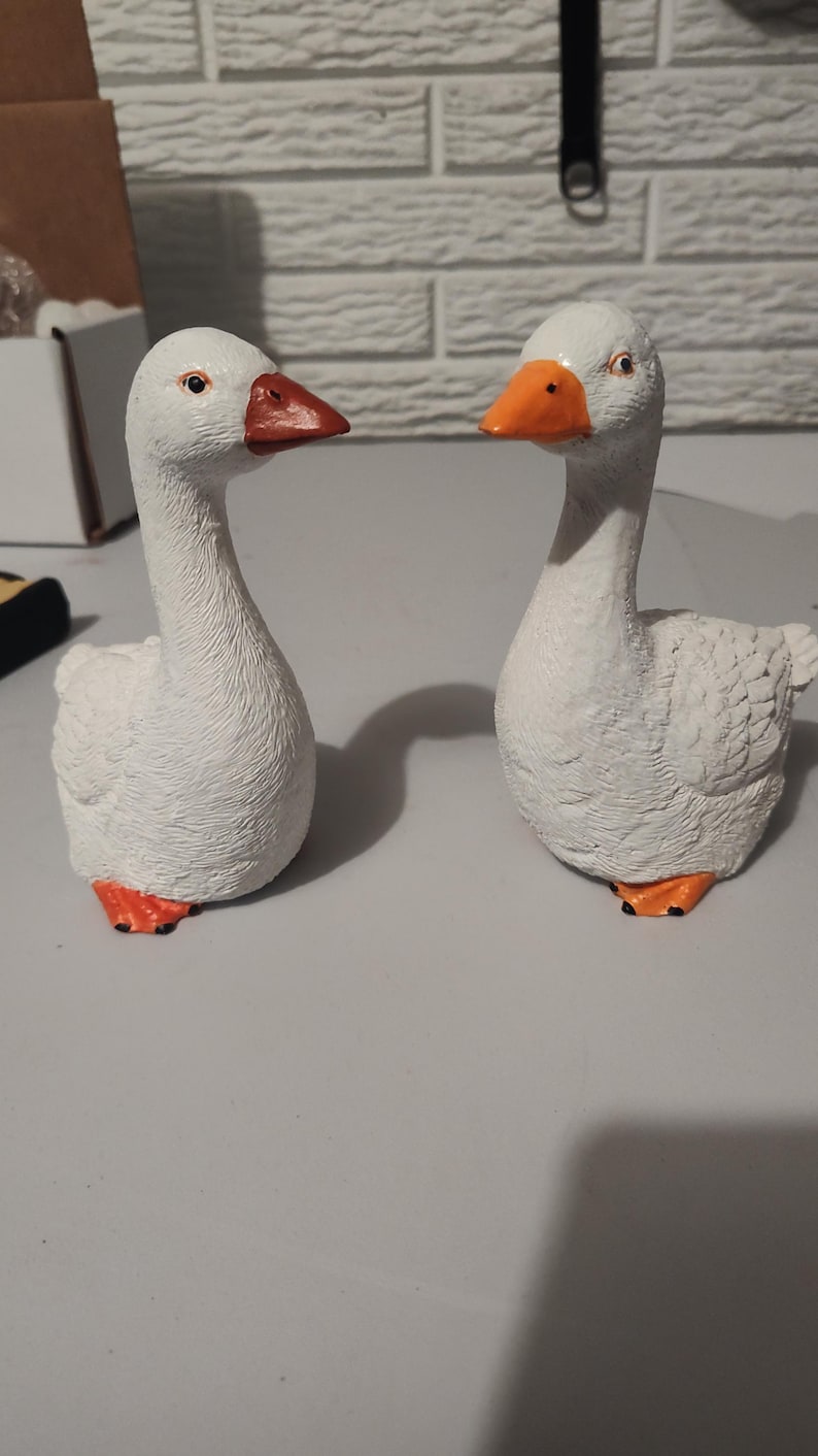 May include: Two white ceramic goose figurines with orange beaks and feet. The geese are facing each other and are positioned on a white surface.