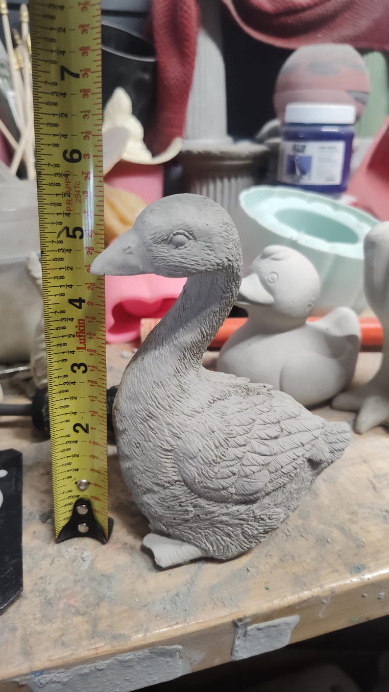 May include: A gray concrete goose figurine measuring approximately 7 inches tall. The goose is sitting with its head turned to the side and its wings folded.