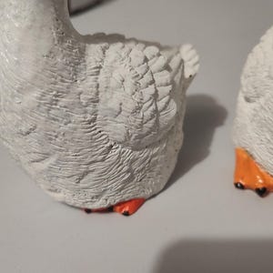May include: Two white ceramic goose figurines with orange feet. The geese are facing away from the camera and have detailed feathers.