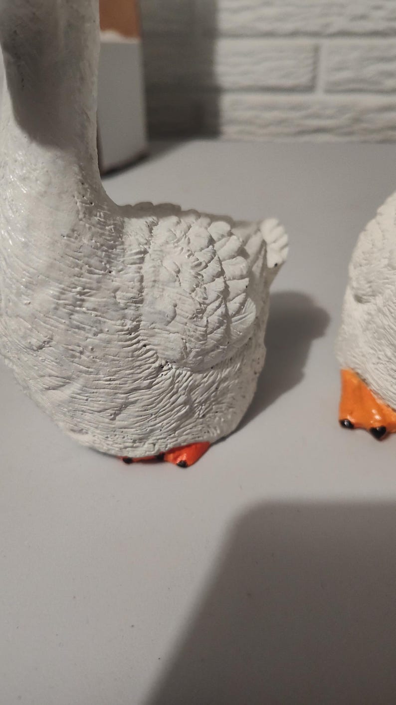 May include: Two white ceramic goose figurines with orange feet. The geese are facing away from the camera and have detailed feathers.