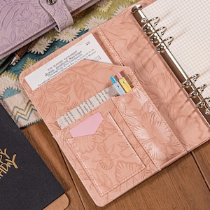 Puede incluir: Carpeta de cuero rosa claro abierta, con relieve floral. La carpeta está abierta, revelando un bolsillo con bolígrafos, una tarjeta rosa y un trozo de papel. El texto "THE BLESSED SACRAMENT" es visible en el papel. Un cuaderno es visible a la derecha.