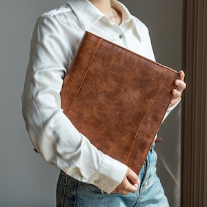 May include: A brown leather portfolio, held by a person in a white shirt and jeans. The portfolio has a textured finish and stitched edges. The design is classic and professional, suitable for business or personal use.