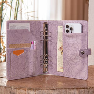 May include: Open lavender-colored binder with a floral embossed design. The binder is open, revealing pockets holding a phone, pens, cards, and papers. A silver metal ring binder mechanism is visible. The binder is on a wooden surface.
