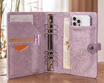 May include: Open lavender-colored binder with a floral embossed design. The binder is open, revealing pockets holding a phone, pens, cards, and papers. A silver metal ring binder mechanism is visible. The binder is on a wooden surface.