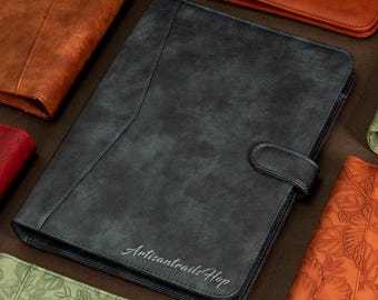 May include: A dark gray leather planner with a flap closure and a silver embossed inscription that reads "ArtisantrailsHop".