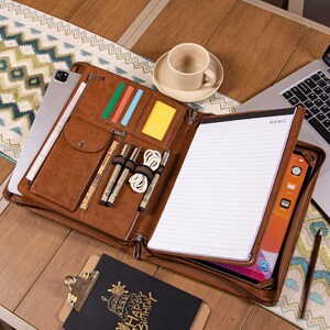 May include: A brown leather portfolio with multiple compartments, including slots for cards, pens, and a tablet. The portfolio is open, revealing a notepad and a tablet. A cup of coffee and a laptop are also visible on the wooden surface.