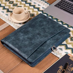 May include: A dark teal leather portfolio with a zipper closure and a flap. The portfolio is on a wooden surface with a patterned runner, a laptop, and a coffee cup.