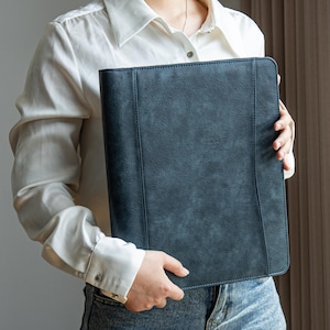 May include: A dark blue leather portfolio held by a person. The portfolio has a textured surface and a vertical seam down the center. The person is wearing a white button-down shirt and blue jeans.