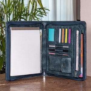 May include: Open, dark blue leather portfolio with multiple compartments. Features a notepad, card slots, pen holders, and a pocket. Includes a zippered closure and a tablet sleeve. The interior is lined with a lighter blue material.