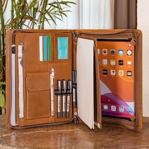 Könnte beinhalten: Geöffnetes braunes Leder-Portfolio mit mehreren Fächern, darunter Kartenfächer, Stifthalter und ein Notizblock. Auf der rechten Seite ist ein iPad zu sehen. Das Portfolio ist mit einem Reißverschluss versehen und liegt auf einer Holzoberfläche.