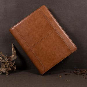 May include: A brown leather portfolio with a textured surface and a flap closure. The portfolio is closed and lying on a brown surface.