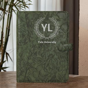 Op de afbeelding: Een olijfgroene folio-omslag met een reliëf bloemenpatroon over het oppervlak. Het midden toont een witte lauwerkransafbeelding die de letters YL omsluit, met de tekst Yale University in het wit direct onder het embleem gedrukt.