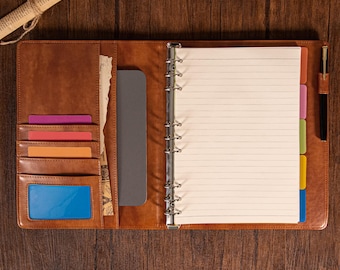 May include: A brown leather-effect binder featuring a six-ring metal mechanism, lined paper, and multiple interior slots for cards. The organiser includes a black pen in a side loop, colourful tab dividers, and a clear window pocket for identification.