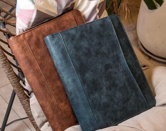 May include: Two leather-look document holders in shades of brown and blue-grey rest on a chair. The brown holder has a warm, earthy tone, while the blue-grey offers a cool contrast. Both feature a textured finish.