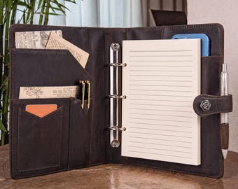 Puede incluir: Una carpeta de cuero marrón oscuro con un mecanismo de tres anillas de metal, un bloc de notas rayado y varios bolsillos interiores. El organizador incluye un portalápices con un bolígrafo plateado, una tarjeta naranja y papeles insertados.