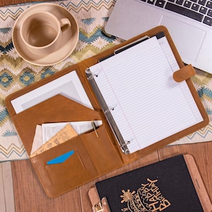 May include: A tan leather portfolio organiser featuring a three-ring binder mechanism, a lined notepad, multiple interior pockets for documents, a pen loop, and a secure strap closure. The interior contains loose papers and a small blue card.