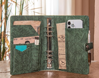 May include: Open green leather binder with floral embossed design. The binder is open, revealing pockets with cards, a phone, and a pen. The binder is sitting on a wooden surface.