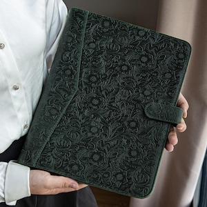 May include: A dark green leather portfolio featuring an embossed floral design. The portfolio has a flap closure with a matching button. The cover is a deep forest green colour, and the floral design is intricate and detailed.