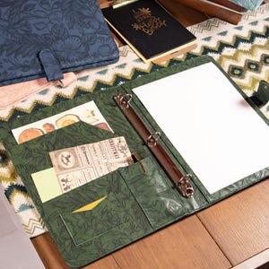 Op de afbeelding: Groene leren map met een bloemmotief, open om een blanco wit vel papier te tonen. De map heeft metalen ringen en vakken met kaarten en een pen. Een blauw leren notitieboek met bloemen en een zwarte "Happy Birthday" kaart zijn ook zichtbaar.