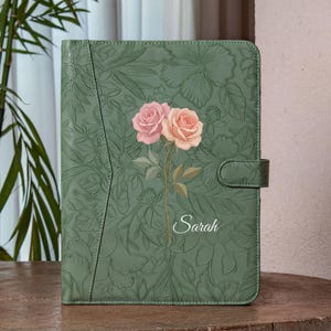 May include: A green leather journal with embossed floral patterns and a clasp closure. The cover features a painted design of two roses in pink and peach, with the name "Sarah" in script.