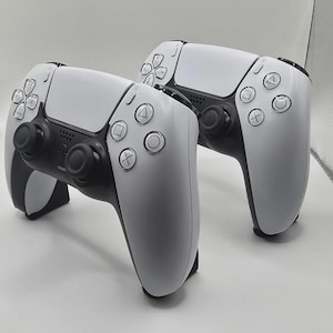May include: Two white and black wireless video game controllers featuring a central touchpad, dual analogue sticks, a directional pad, and four action buttons with triangle, circle, cross, and square symbols. The controllers have a sleek, ergonomic design.