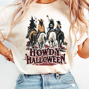 Puede incluir: Camiseta color crema con un gr&aacute;fico de cuatro jinetes a caballo y el texto "HOWDY HALLOWEEN" en una fuente desgastada de estilo occidental. Los jinetes visten disfraces de Halloween. La camiseta se combina con vaqueros de mezclilla de lavado claro.