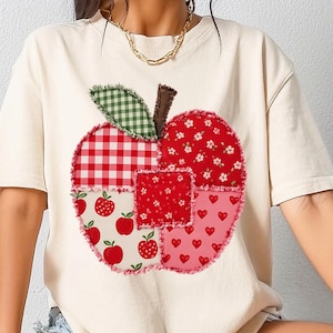 May include: A cream-colored short-sleeve t-shirt featuring a large, central apple-shaped graphic. The apple is composed of various red and white patterned fabric patches, including gingham, floral, and heart prints, with a green checkered leaf.