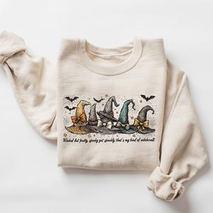 May include: Cream-colored sweatshirt featuring a graphic of five witch hats in various colors, with bats and stars. The text "Wicked but pretty, spooky yet sparkly, that's my kind of witchcraft" is printed below the hats.
