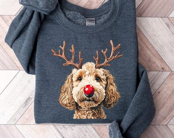 May include: A dark gray sweatshirt featuring a festive illustration of a dog with reindeer antlers and a red nose. The dog has brown fur and a white face. The sweatshirt is folded on a wooden surface.