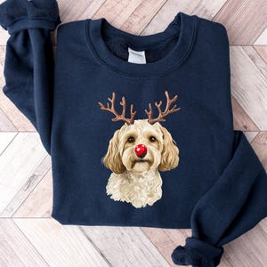 May include: Navy blue sweatshirt featuring a dog with reindeer antlers and a red nose. The dog has light brown fur and is centered on the front of the sweatshirt. The sweatshirt has a crew neck and long sleeves.
