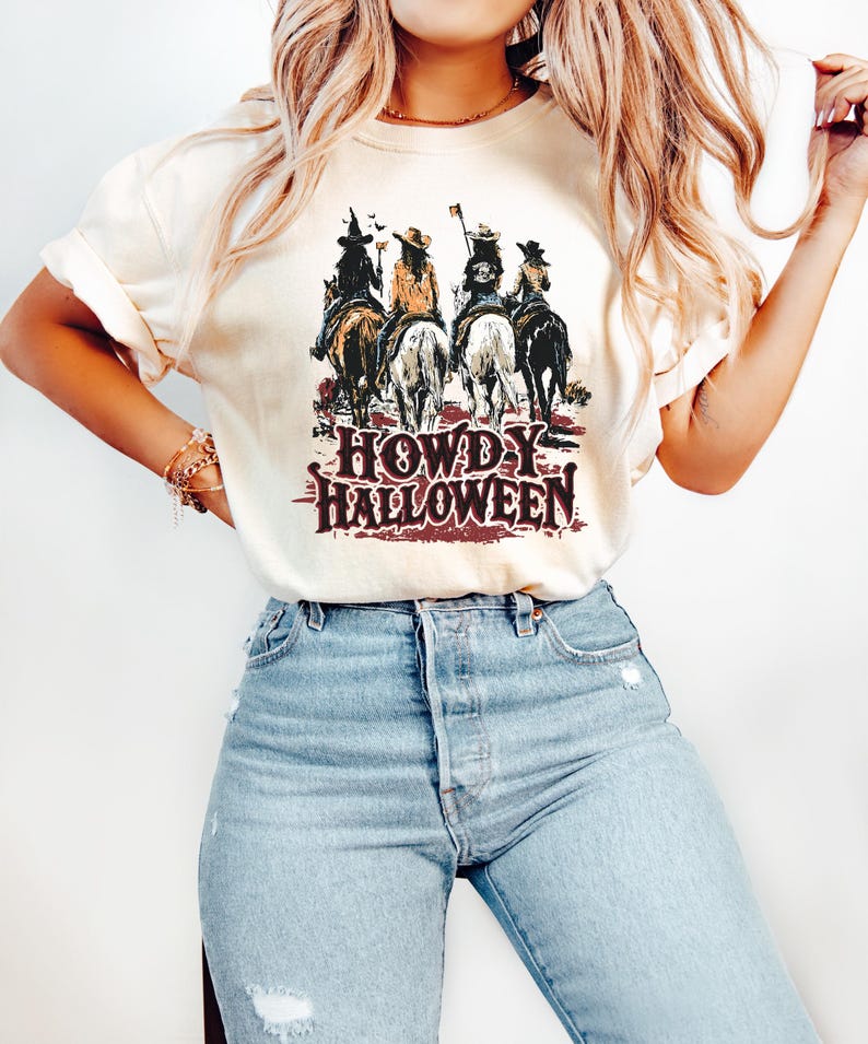 Puede incluir: Camiseta color crema con un gr&aacute;fico de cuatro jinetes a caballo y el texto "HOWDY HALLOWEEN" en una fuente desgastada de estilo occidental. Los jinetes visten disfraces de Halloween. La camiseta se combina con vaqueros de mezclilla de lavado claro.