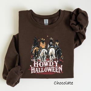 Puede incluir: Sudadera de color chocolate con un gr&aacute;fico de cuatro vaqueros a caballo y el texto "HOWDY HALLOWEEN". La sudadera tiene mangas largas y cuello redondo. La palabra "Chocolate" est&aacute; impresa en la parte inferior.