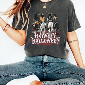 Puede incluir: Camiseta gris oscuro con un gr&aacute;fico de vaqueros a caballo, con el texto "HOWDY HALLOWEEN" en rojo y blanco. La camiseta se lleva con vaqueros azules y zapatillas blancas. Se ve un collar y una pulsera dorados.