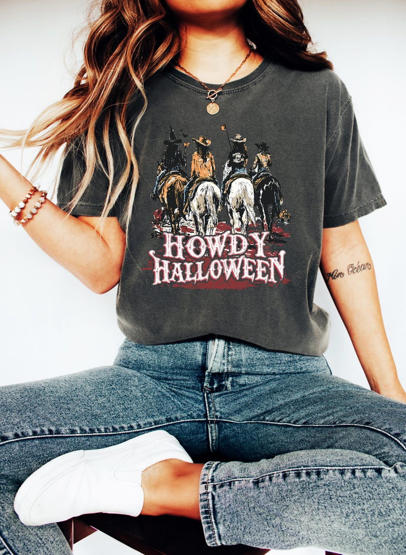 Puede incluir: Camiseta gris oscuro con un gr&aacute;fico de vaqueros a caballo, con el texto "HOWDY HALLOWEEN" en rojo y blanco. La camiseta se lleva con vaqueros azules y zapatillas blancas. Se ve un collar y una pulsera dorados.