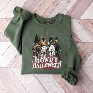 Puede incluir: Sudadera verde oliva con un gr&aacute;fico de cuatro jinetes a caballo y el texto "HOWDY HALLOWEEN". El dise&ntilde;o tiene un tema del oeste, con un fondo de puesta de sol. Una prenda festiva con tem&aacute;tica de Halloween.
