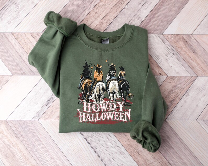Puede incluir: Sudadera verde oliva con un gr&aacute;fico de cuatro jinetes a caballo y el texto "HOWDY HALLOWEEN". El dise&ntilde;o tiene un tema del oeste, con un fondo de puesta de sol. Una prenda festiva con tem&aacute;tica de Halloween.