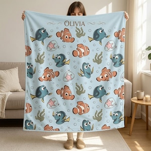 May include: A light blue fleece blanket featuring a repeating pattern of orange clownfish, blue tang fish, pink starfish, and green seaweed. The name OLIVIA is printed in dark brown text at the top center, surrounded by small white bubbles.