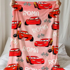 Puede incluir: Una manta rosa con un patr&oacute;n repetido de coches de carreras rojos de dibujos animados, banderas a cuadros y la palabra "ROMA" en blanco. La manta se muestra sobre un fondo blanco.