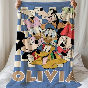 Pode incluir: Uma manta aos quadrados azuis e brancos com um gráfico colorido do Mickey, Minnie, Pato Donald, Margarida, Pateta e Pluto. O nome OLIVIA está impresso em letras maiúsculas grandes, a negrito e de cor clara na parte inferior.